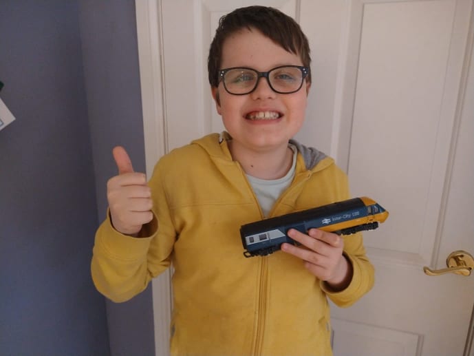 A boy stands facing the camera, he is wearing a bright yellow hoodie jumper and a big smile on his face. the boy is called Alex and he has a model train in one hand and a thumbs up in his other hand.