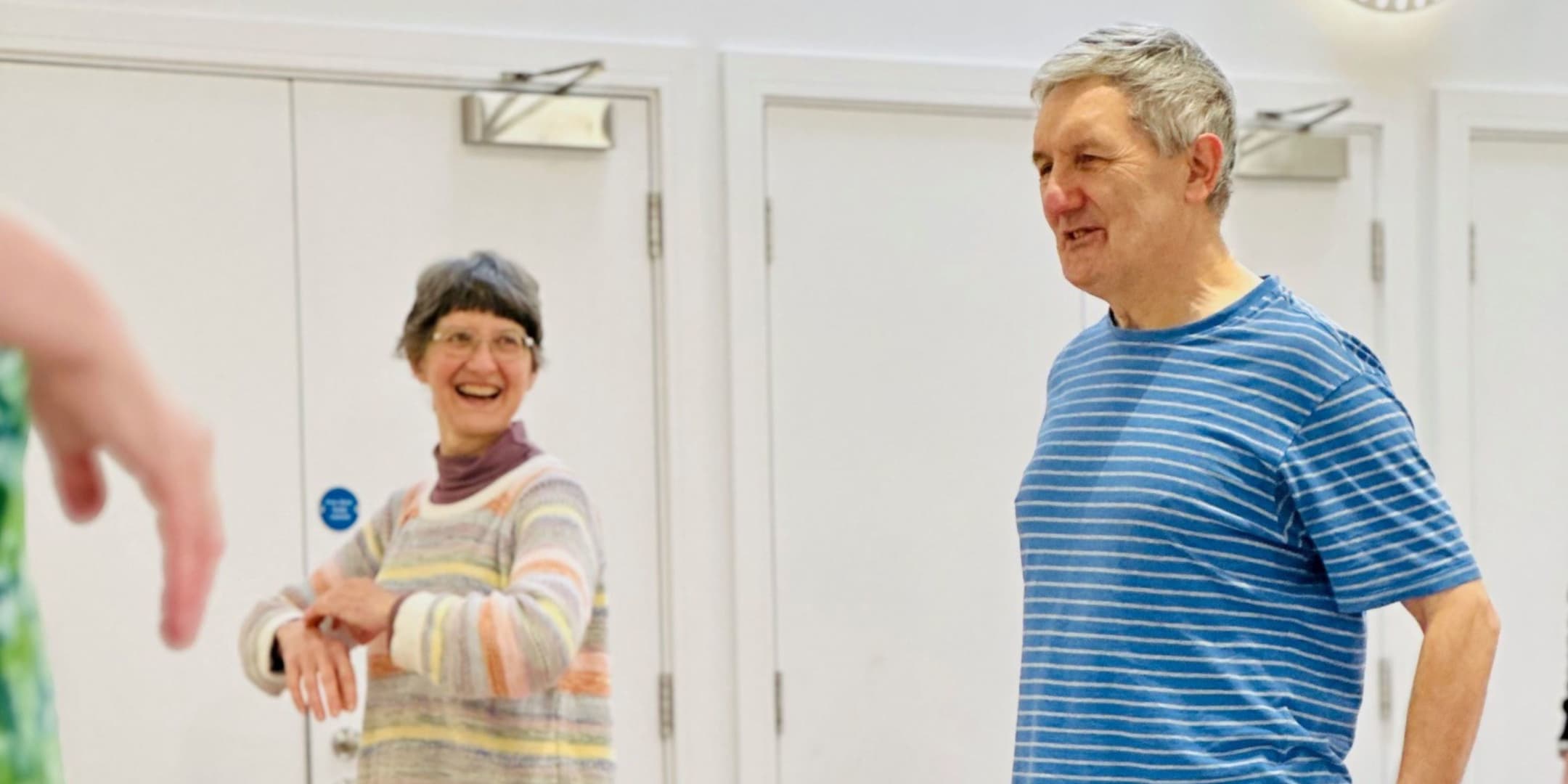 A lady wearing a bright striped jumper looks to the man on her left and smiles - she is holding her arms up and doing exercises. He is wearing a blue T shirt and saying something which is making her laugh. They are in white painted room and the pictu