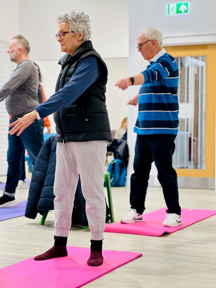 Three people dressed in active wear are standing on bright pink yoga mats, they are turning to the right and stretching out their arms as they do pilates exercises.
