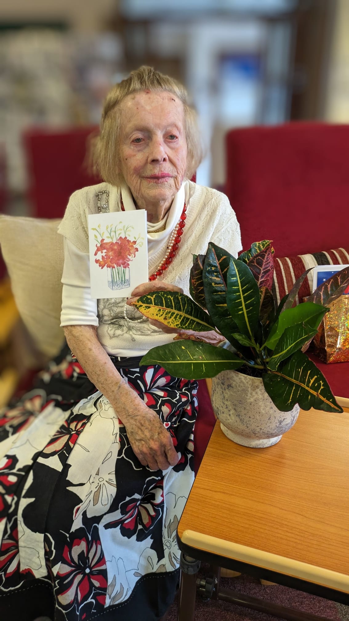 Pictured at her 100th birthday party Kaisu celebrates with Cam Sight friends and volunteers. She smiles for the camera with a card and plant.