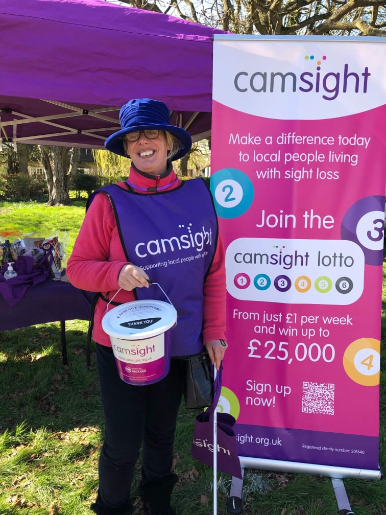 Image of Sue Lambley supporting us at one of our fundraising events holding the Cam Sight bucket with our information banner behind promoting the Lotto Scheme.